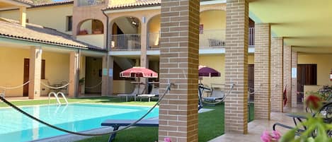 Ett poolområde med en klarblå pool, omgivet av en innergård i tegel och en grön gräsmatta. Här finns solstolar och parasoller, och i bakgrunden syns en byggnad med flera balkonger.