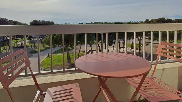 A balcony with a table and two chairs, overlooking a park with trees and a road.