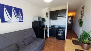 A compact living space with a grey sofa, a small kitchenette, a washing machine, and a dining area with a table and chairs.