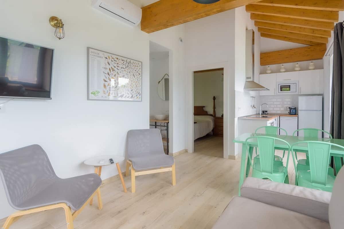 A modern living room with a flat-screen TV, two grey chairs, a small table, and a green dining set.