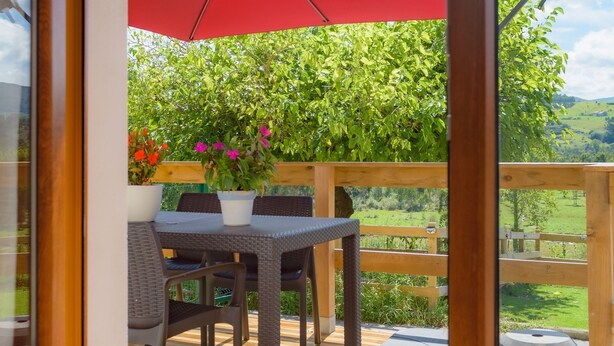 A wooden deck with a table and chairs, a red umbrella, and a potted plant with pink flowers.