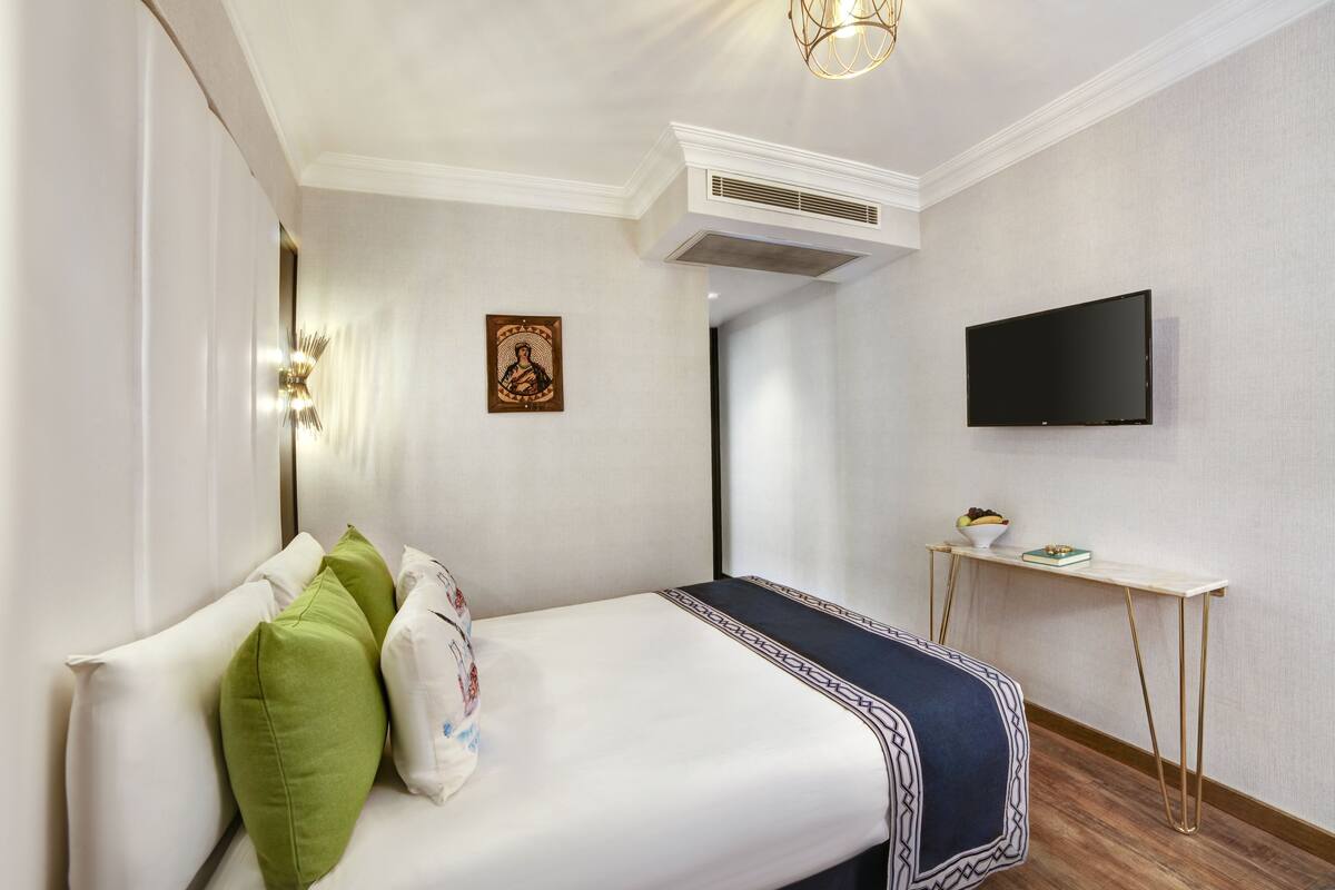 A hotel room with a bed, pillows, a bedside table with a bowl, a flat-screen TV, and a wall-mounted air conditioner.