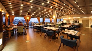 Un ristorante con diversi tavoli e sedie, pavimento in legno e vista sulla città attraverso ampie finestre.