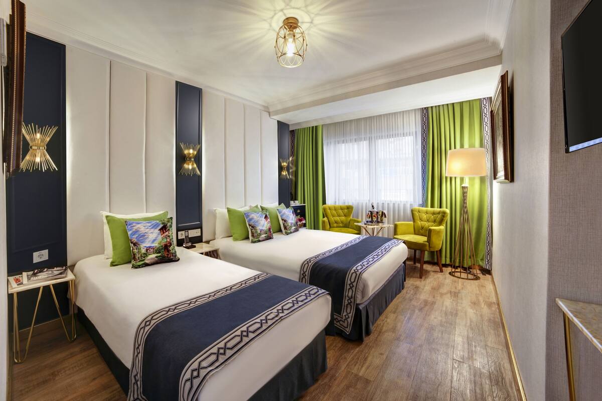 A hotel room with two beds, a green chair, a small table, and a TV on the wall.