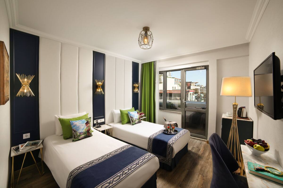 A hotel room with two beds, a desk, a chair, a TV, and a window with a view of buildings.