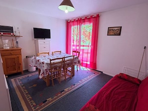 A room with a dining table, chairs, a TV, a cabinet, a trash bin, and a window with red curtains.