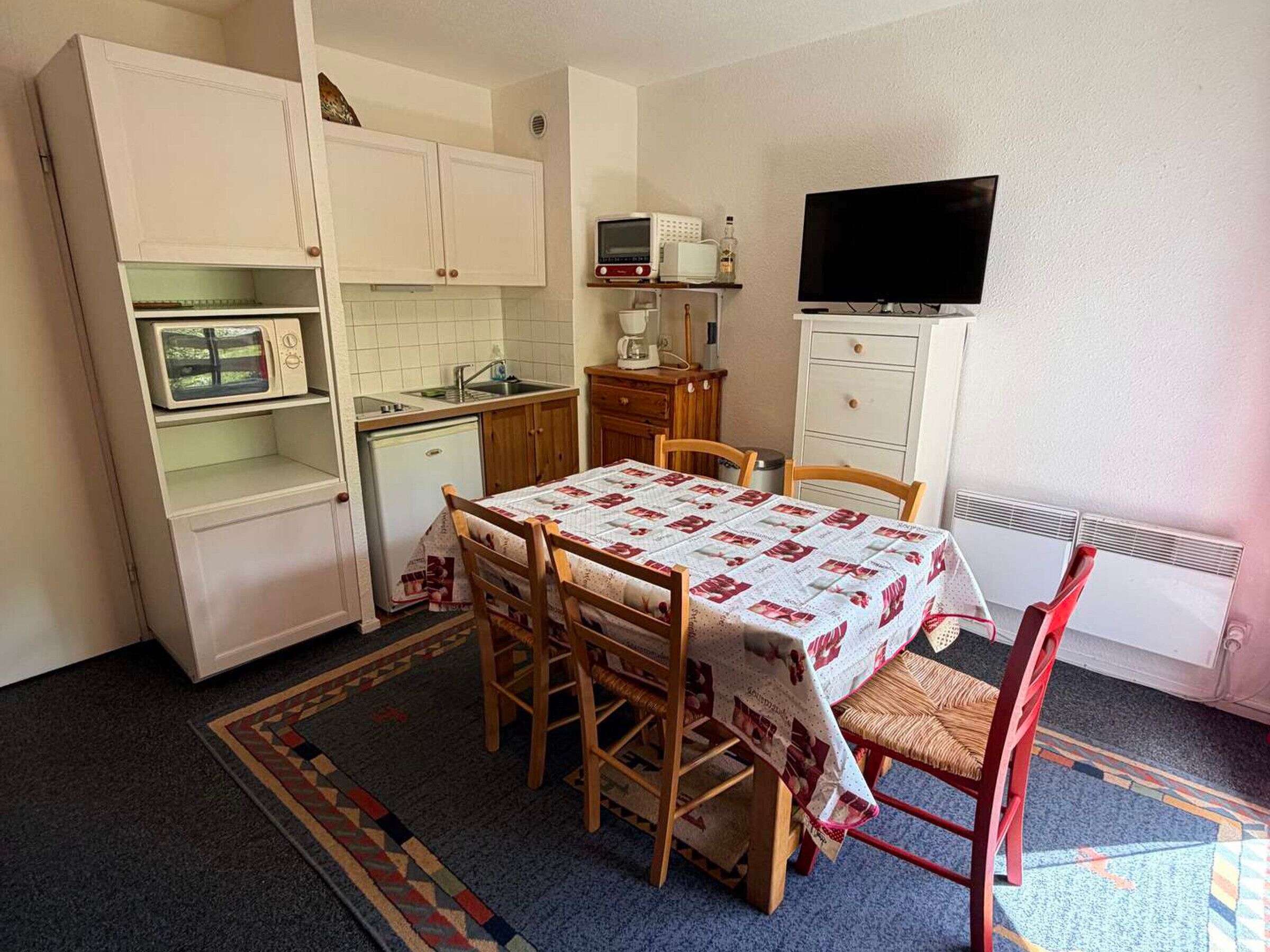 Une cuisine compacte équipée d’un four à micro-ondes, d’un évier et d’un petit réfrigérateur. On y trouve une table à manger avec des chaises, ainsi qu’un téléviseur fixé au mur.