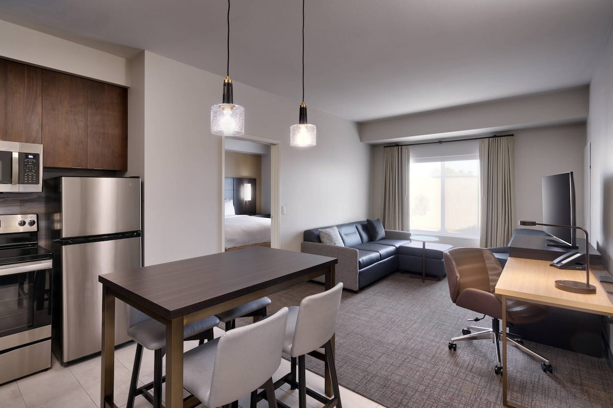 A modern hotel room with a kitchenette, a dining area, a sofa, a desk, and a TV.