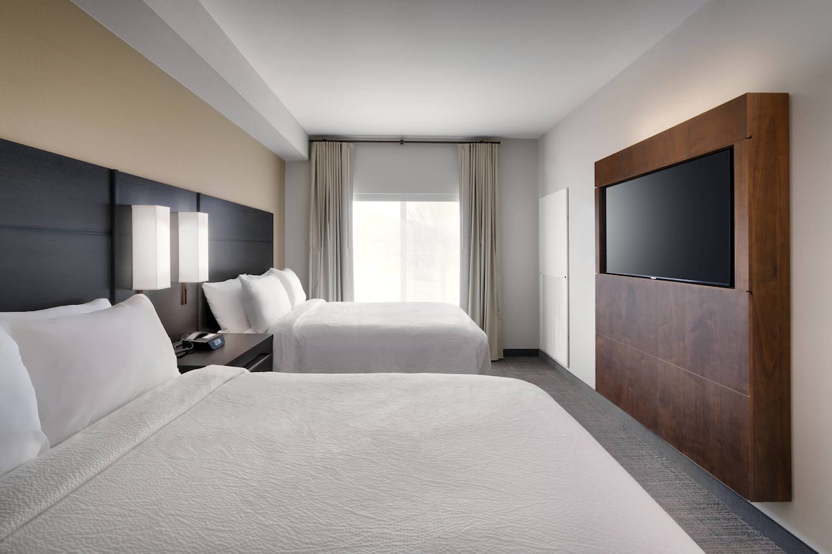 A hotel room with two beds, a flat-screen TV, a desk with a lamp, and a large window with curtains.