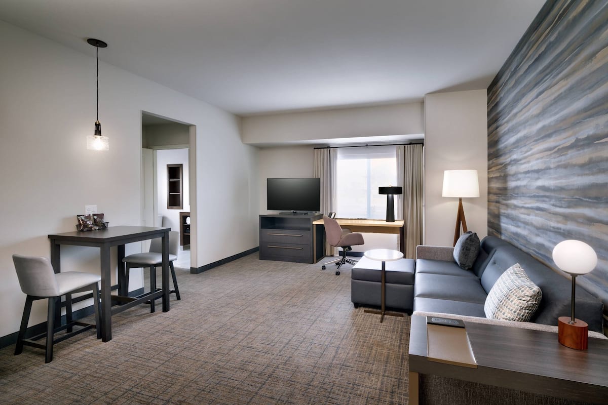 A modern hotel room with a dining area, a sofa, a TV, and a large abstract wall art piece.