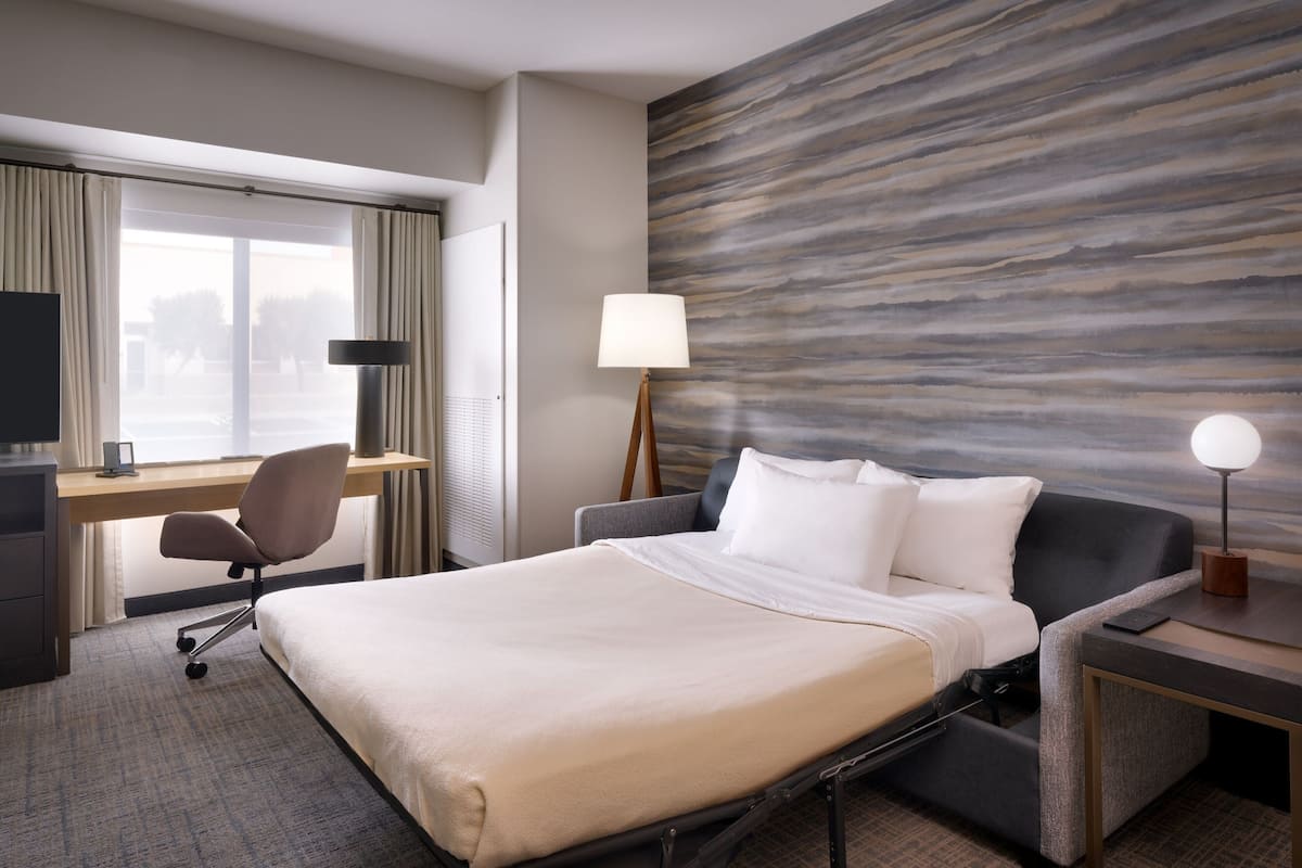 A hotel room with a bed, desk, chair, lamp, and a wall with a wood-like texture.