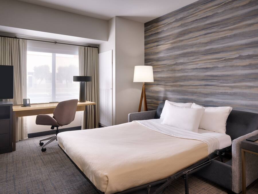 A hotel room with a bed, desk, chair, lamp, and a wall with a wood-like texture.