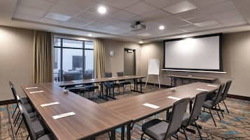 Una sala de conferencias con una larga mesa, sillas, un proyector, una pantalla y vistas de la ciudad a través de amplios ventanales.