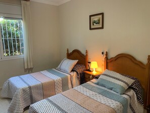 Une chambre avec deux lits simples, une table de chevet avec une lampe, un tableau encadré au mur et une fenêtre avec des rideaux.