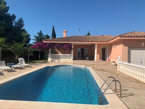 Une piscine à l’eau d’un bleu cristallin, entourée d’une terrasse carrelée. On y trouve des chaises longues et un espace patio couvert avec une table et des chaises.