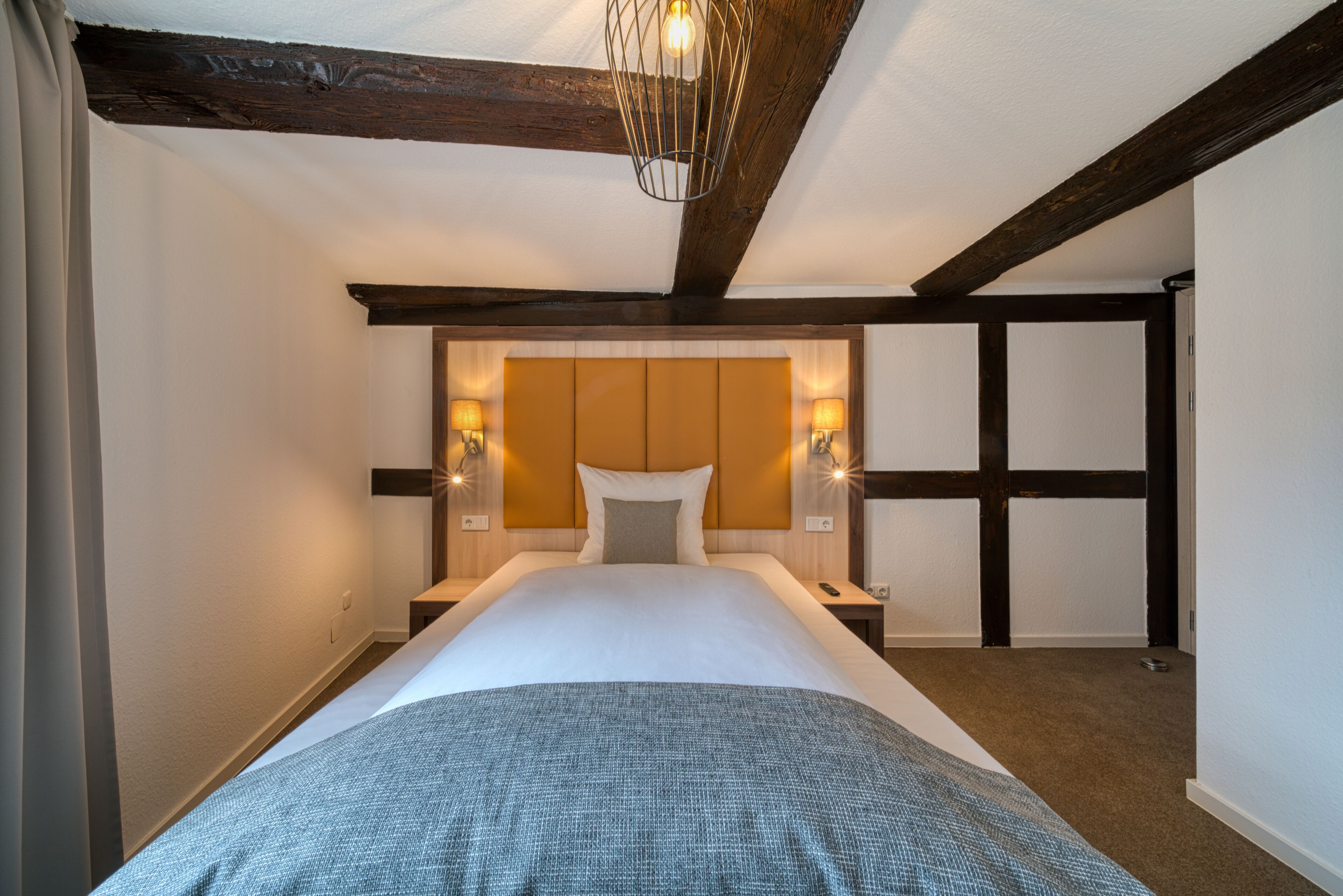 A hotel room with a large bed, wooden beams, a headboard with wall sconces, and a hanging light fixture.