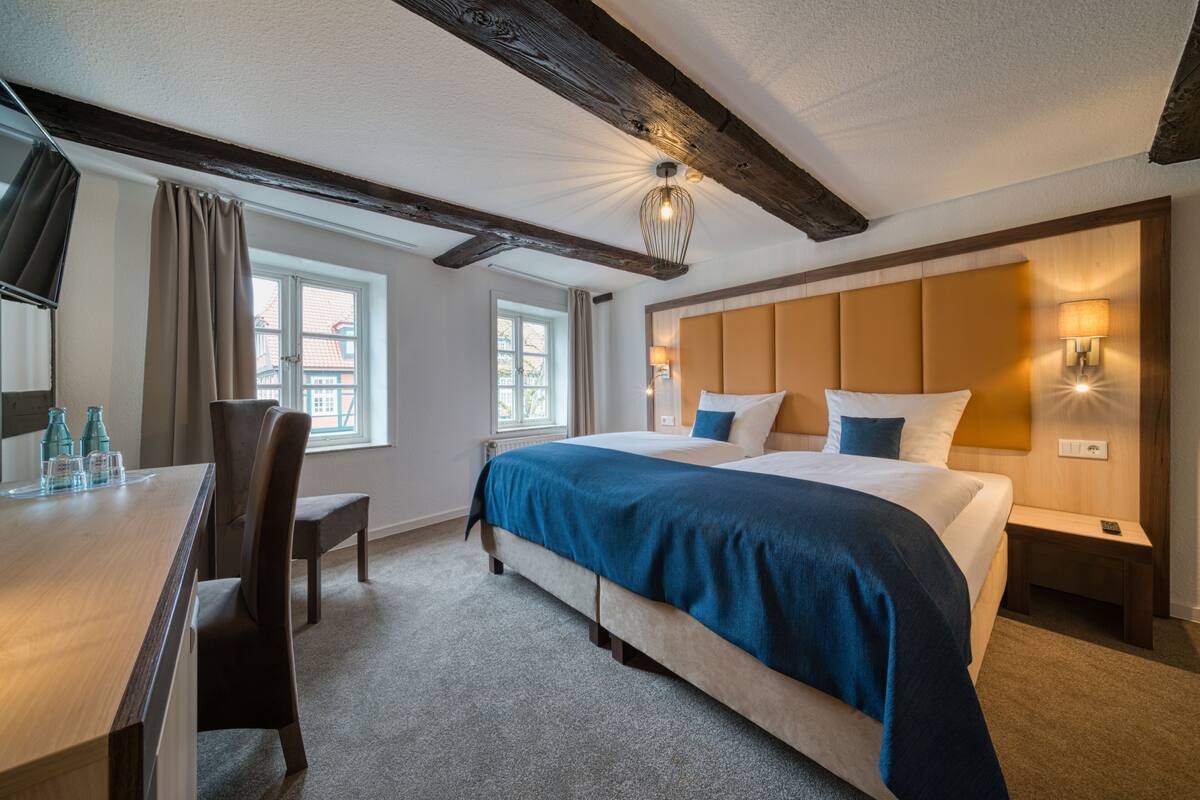 A hotel room with a large bed, a desk, two chairs, a window, and a wooden ceiling beam.