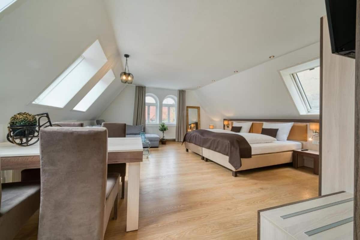 A modern, well-lit room with a loft-style ceiling, a bed, a desk, and a dining area.