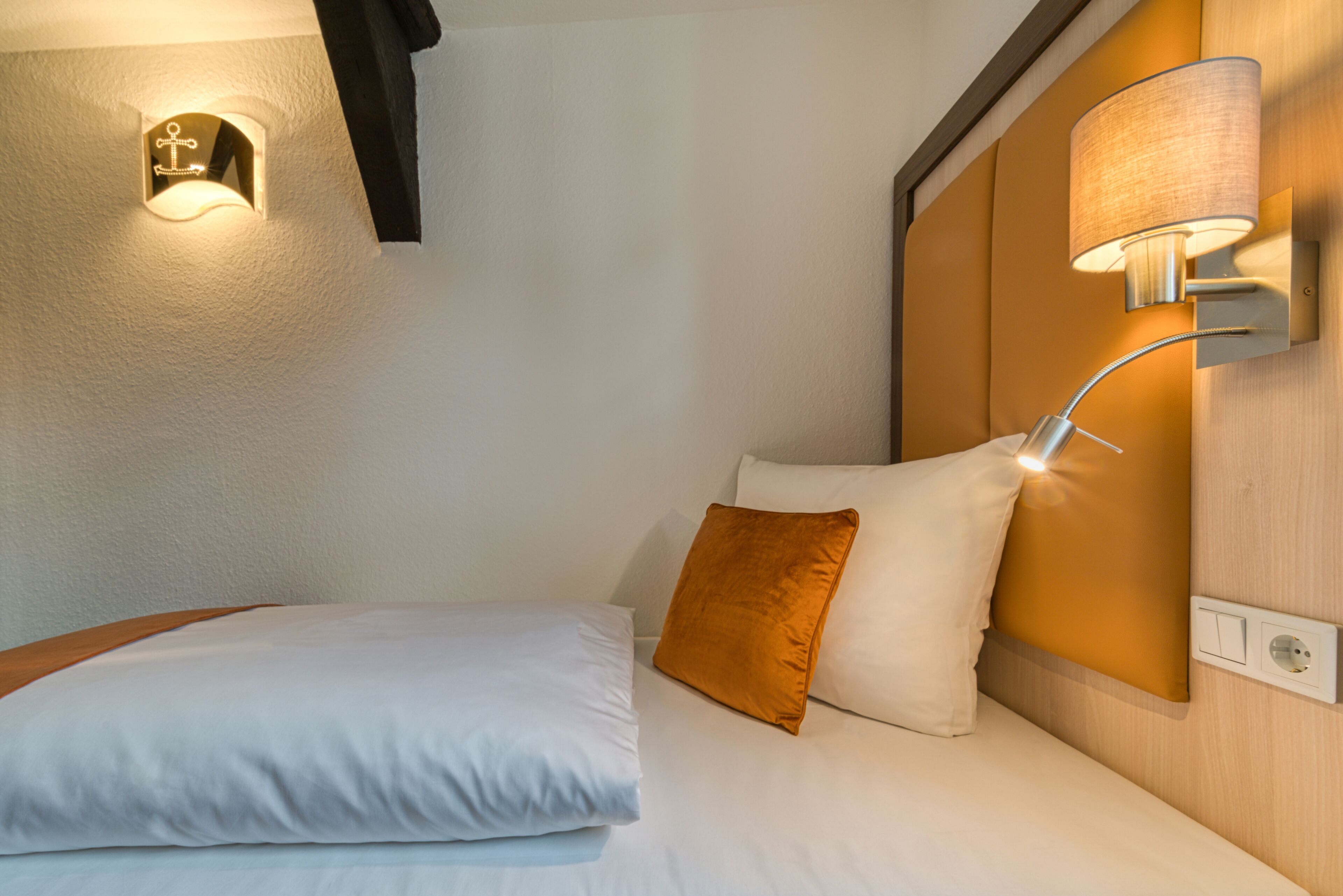 Quarto de hotel com uma cama bem arrumada, cabeceira com luminária e luminária fixada na parede.