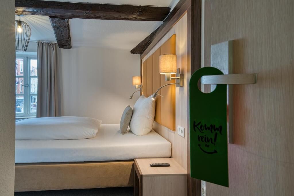 A hotel room with a bed, a nightstand, a wall-mounted lamp, and a green sign with text.