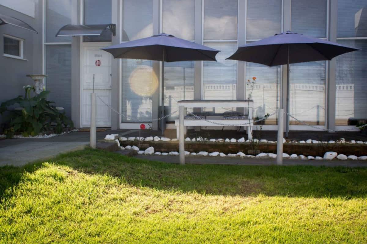 Un patio con dos sombrillas, una mesa y una silla. Se observa un edificio con ventanas amplias al fondo.