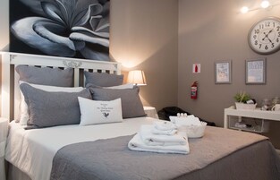 Cama bem arrumada com roupa de cama cinza, luminária de cabeceira, relógio e placa de boas-vindas emoldurada.