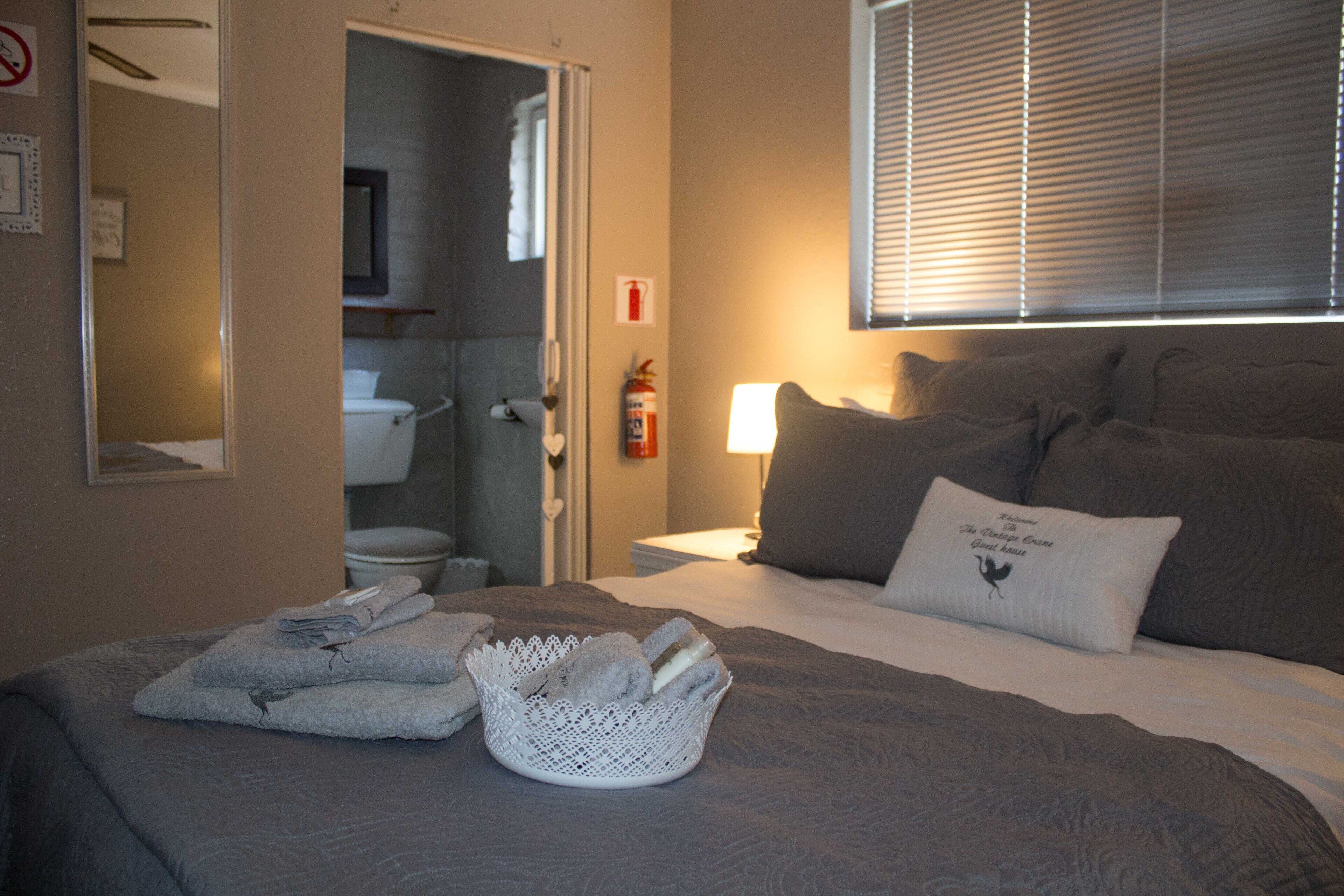 A hotel room with a bed, a bedside table, a lamp, a window with blinds, a bathroom visible through an open door, and a fire extinguisher on the wall.