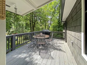 A covered outdoor deck with a round table and chairs, surrounded by a wooden railing and a view of a wooded area. - Secluded, Stylish & Completely Reimagined | Azalea at Highlands Falls (Highlands)