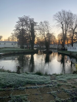 A serene lakeside view with a small pond, surrounded by trees and a row of mobile homes. - Kellysholidayhomes, Atlas, 3 bedroom Caravan, surrounded by nature (Weeley)