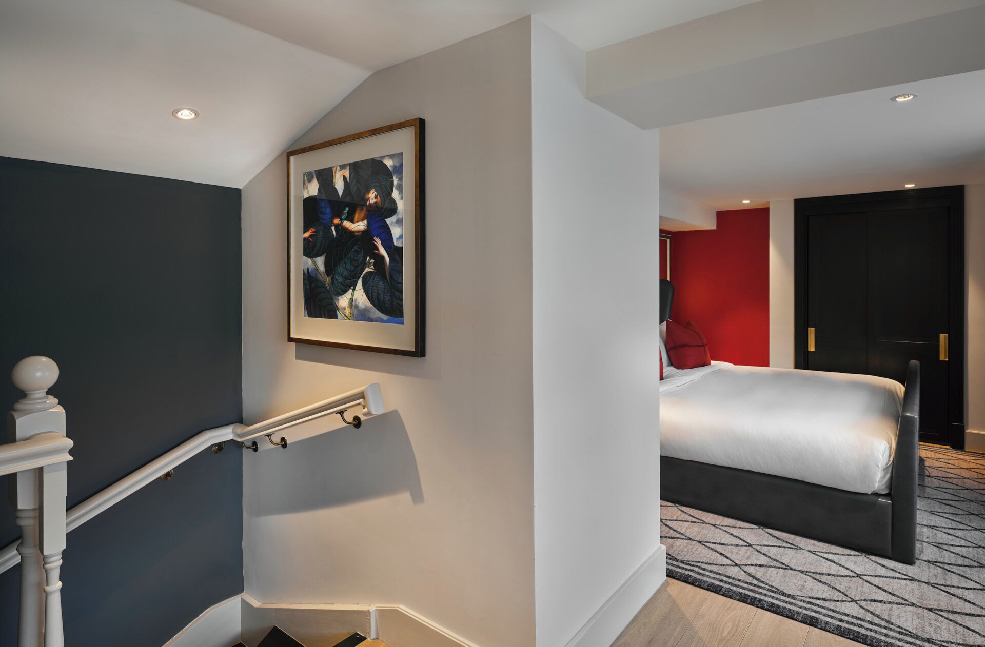 A hotel room with a bed, a staircase with a handrail, a painting on the wall, and a red accent wall.