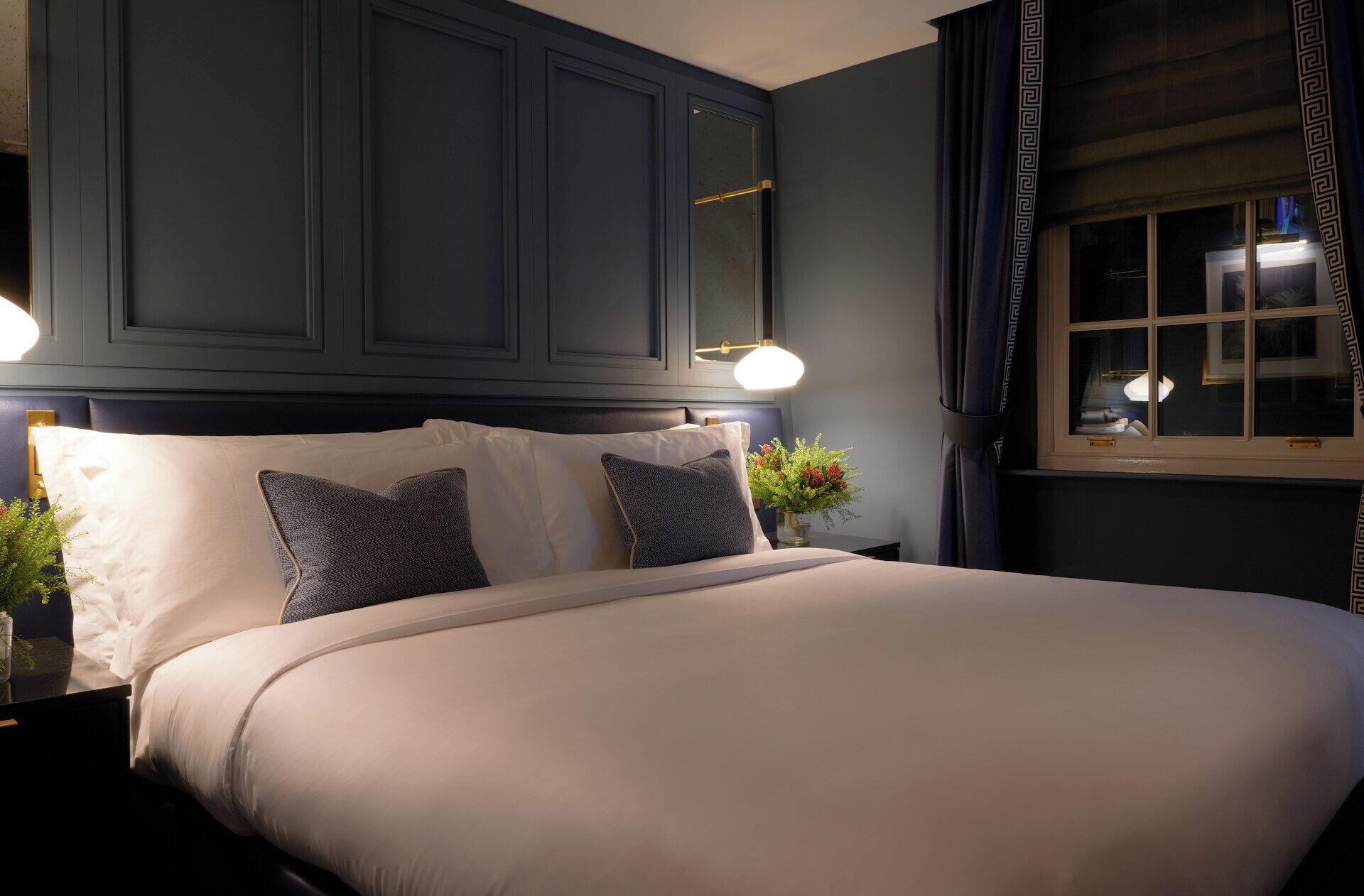 Una cama bien hecha con sábanas blancas y dos almohadas, una mesita de noche con lámpara, y una ventana con cortinas.