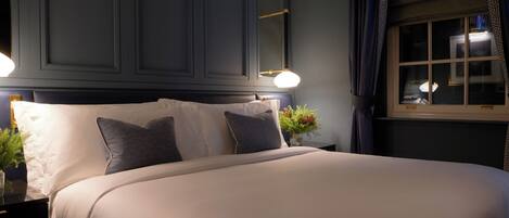 A neatly made bed with white linens and two pillows, a bedside table with a lamp, and a window with curtains.