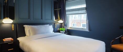 A hotel room with a large bed, a nightstand with a lamp, a window with curtains, and a view of a building outside.