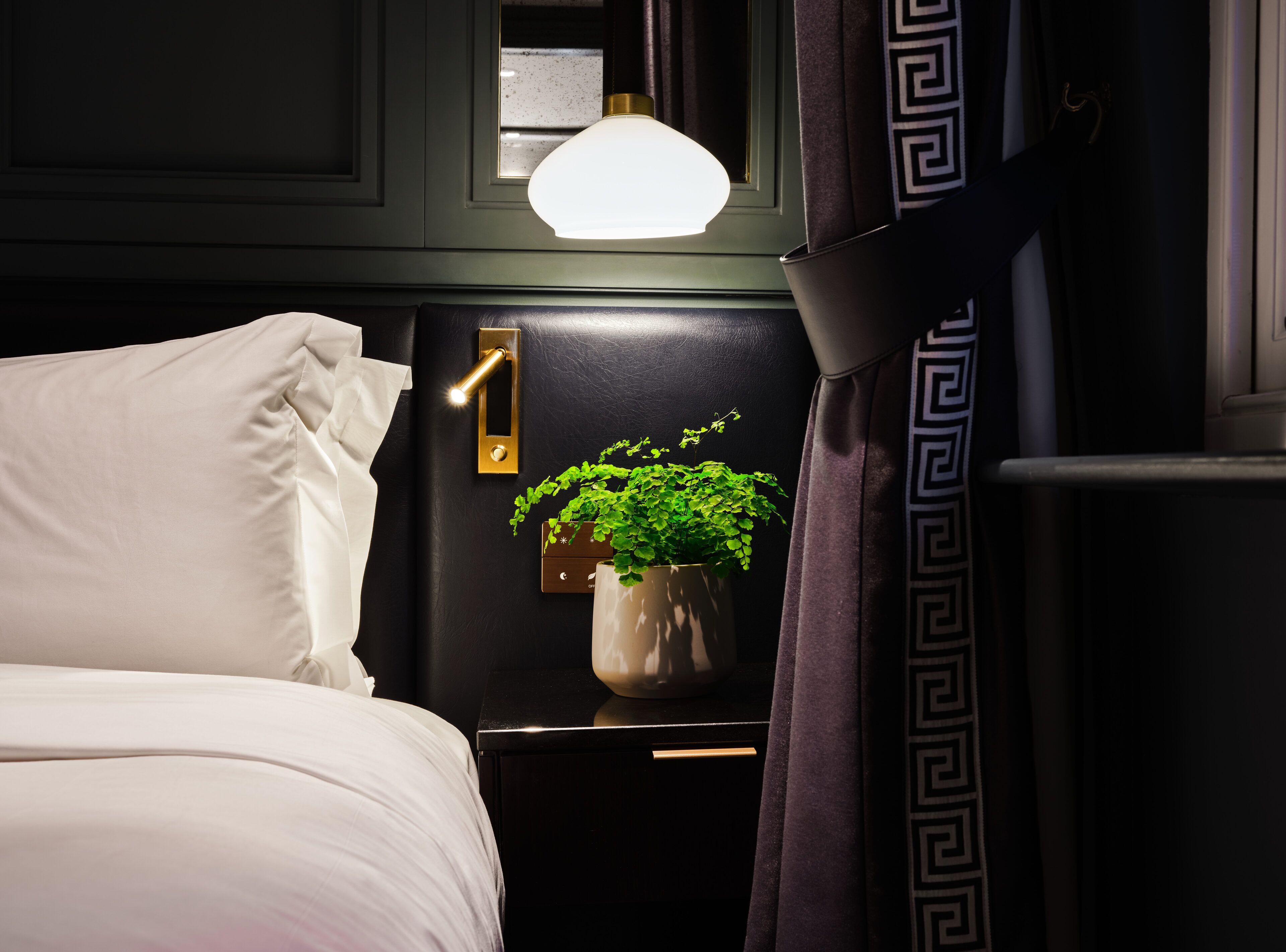 A hotel room with a bed, a nightstand with a lamp, a potted plant, and a curtain with a geometric pattern.
