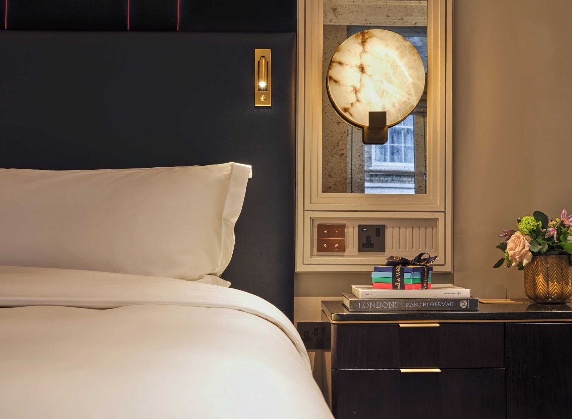Un lit bien fait, avec une tête de lit, une table de chevet avec une lampe, une fenêtre donnant sur un beau paysage et une petite bibliothèque remplie de livres.