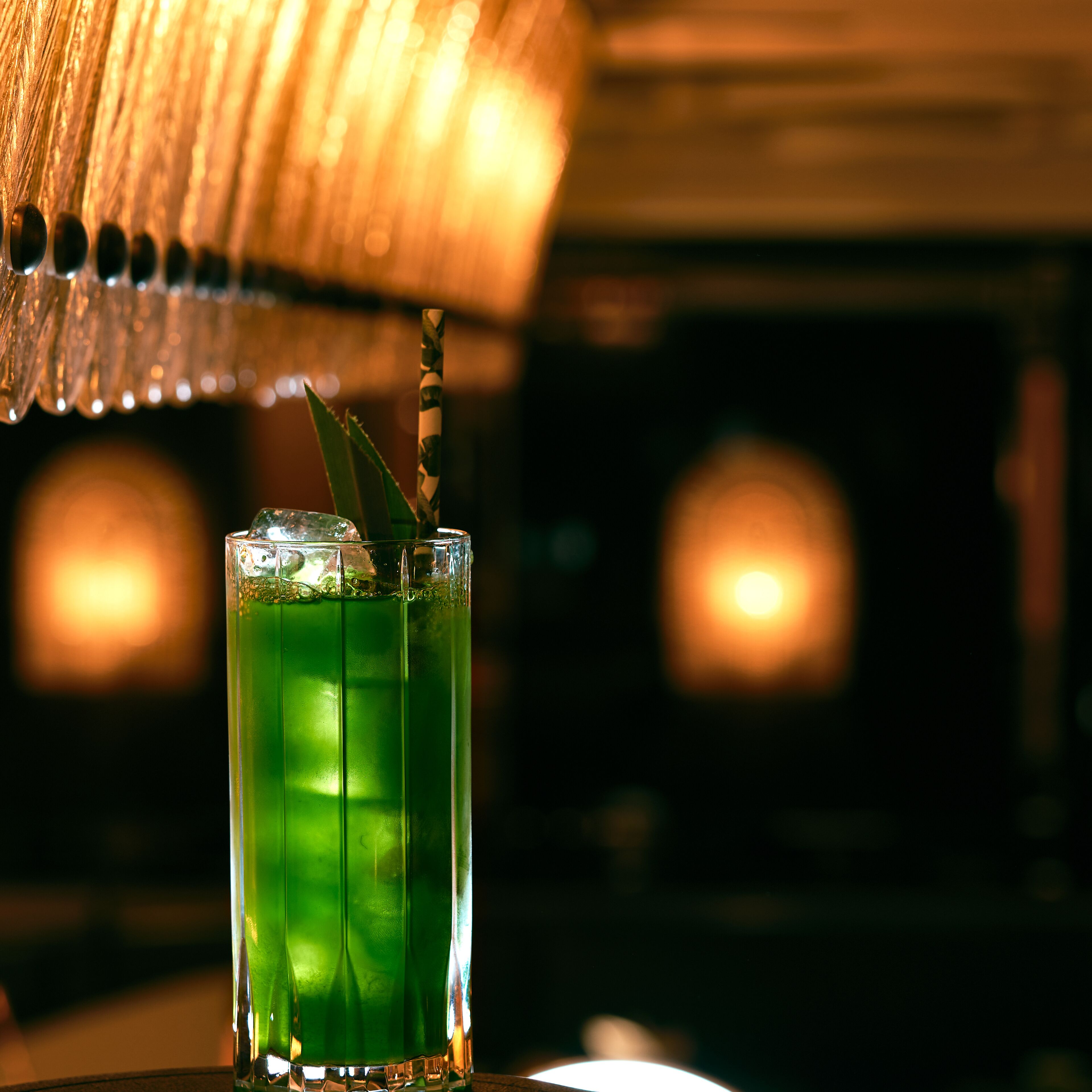 Un vaso con bebida verde, un cubito de hielo y una pajita, sobre una mesa con fondo desenfocado.