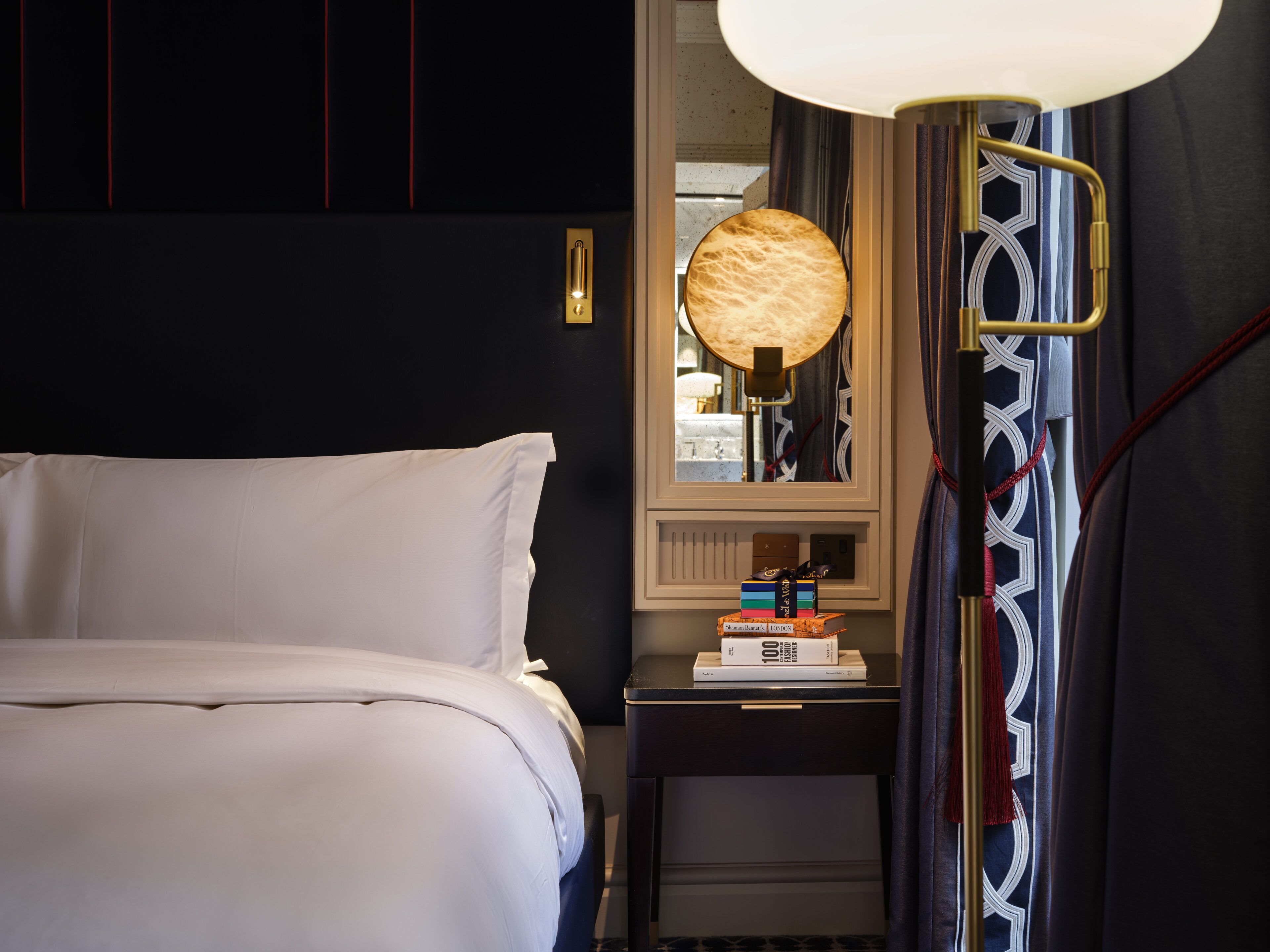 A neatly made bed with white linens, a bedside table with a lamp, and a mirror reflecting the room.