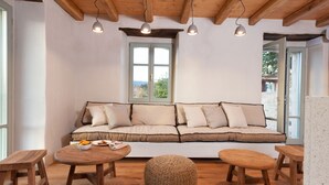 Ein gemütliches Wohnzimmer mit Holzbalkendecke, einem beigefarbenen Sofa mit Kissen, einem runden gewebten Hocker und zwei Holztischen.