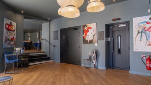 A spacious lobby with a wooden floor, black doors, and a staircase leading to another level. There are blue chairs and a small table with a vase of flowers. -  Boutique Hôtel Nice Côte d'Azur (Nice)
