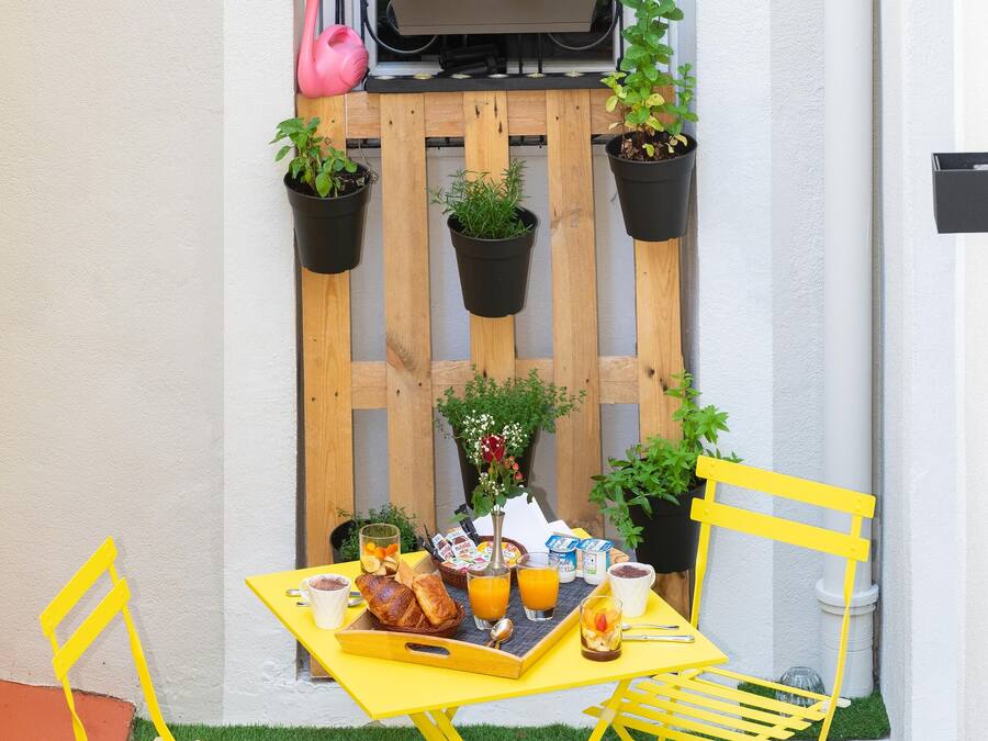 Un pequeño espacio exterior para comer con una mesa y sillas amarillas, rodeado de plantas en macetas y una valla de madera.