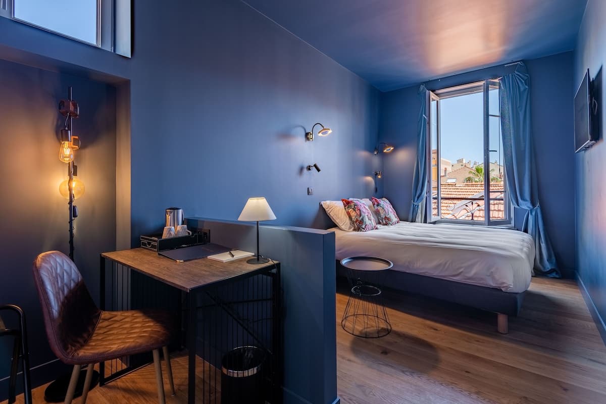 A compact hotel room with a bed, a desk with a chair, a small table, and a window with curtains.