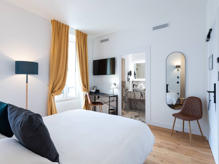 Una habitación de hotel moderna con una cama grande, un escritorio, una silla, un televisor, un espejo y un baño visible a través de una puerta abierta.