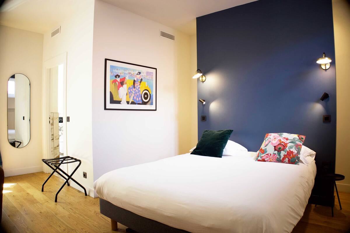 A bedroom with a bed, a mirror, a painting, and wall-mounted lights.