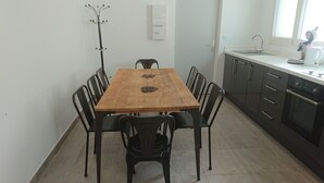 A dining area with a wooden table and black chairs, a coat rack, a wall-mounted electrical panel, a kitchen with a sink and stove, and a window with blinds.