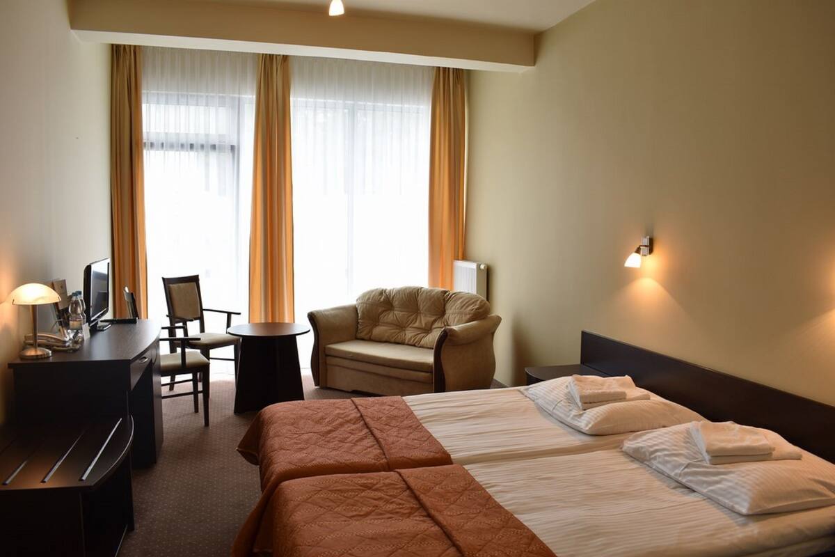 Habitación de hotel con dos camas, un escritorio con lámpara, una silla, un sofá y un ventanal con cortinas.