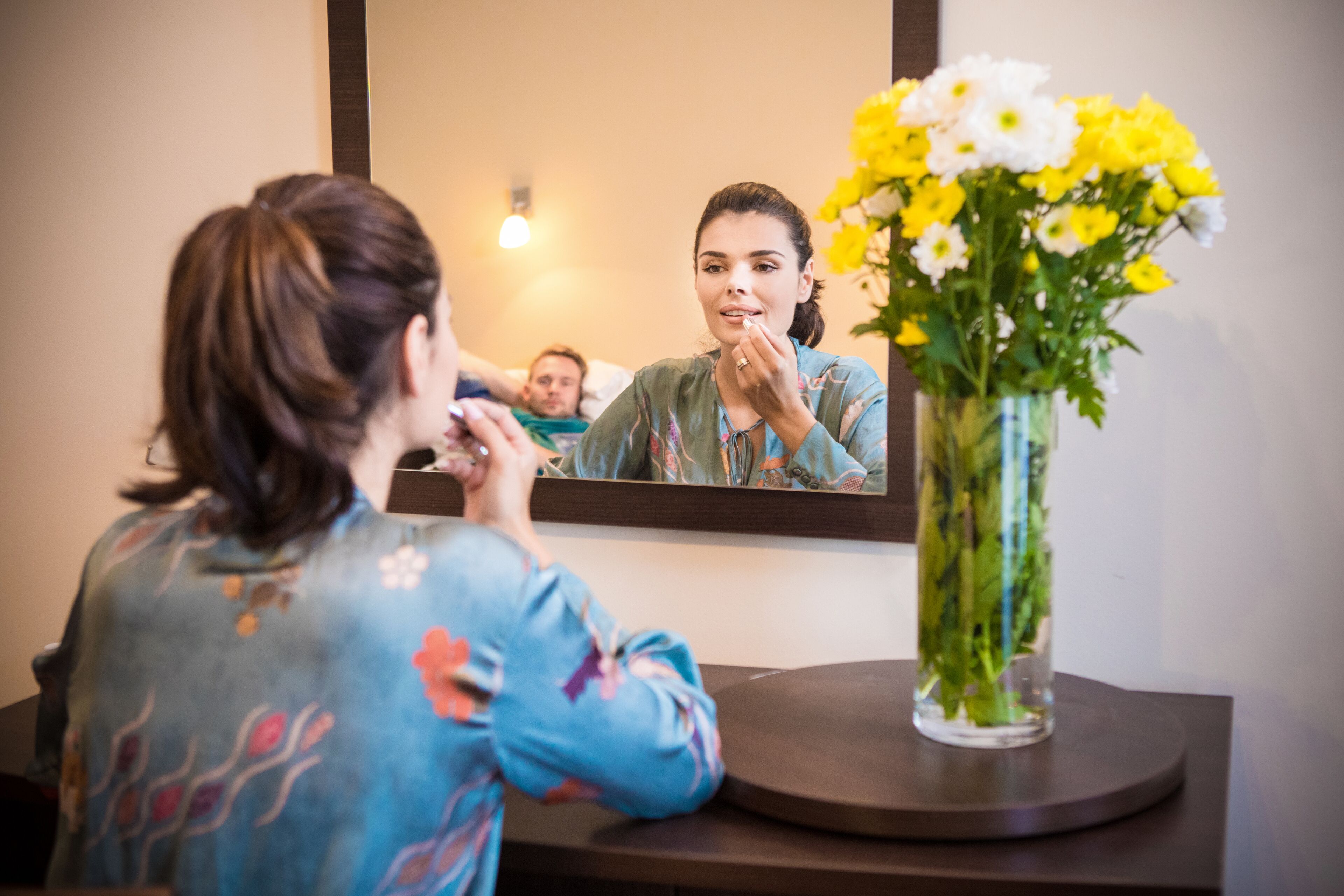 A woman sitting at a table with a mirror, a vase of flowers, and a man in the background.