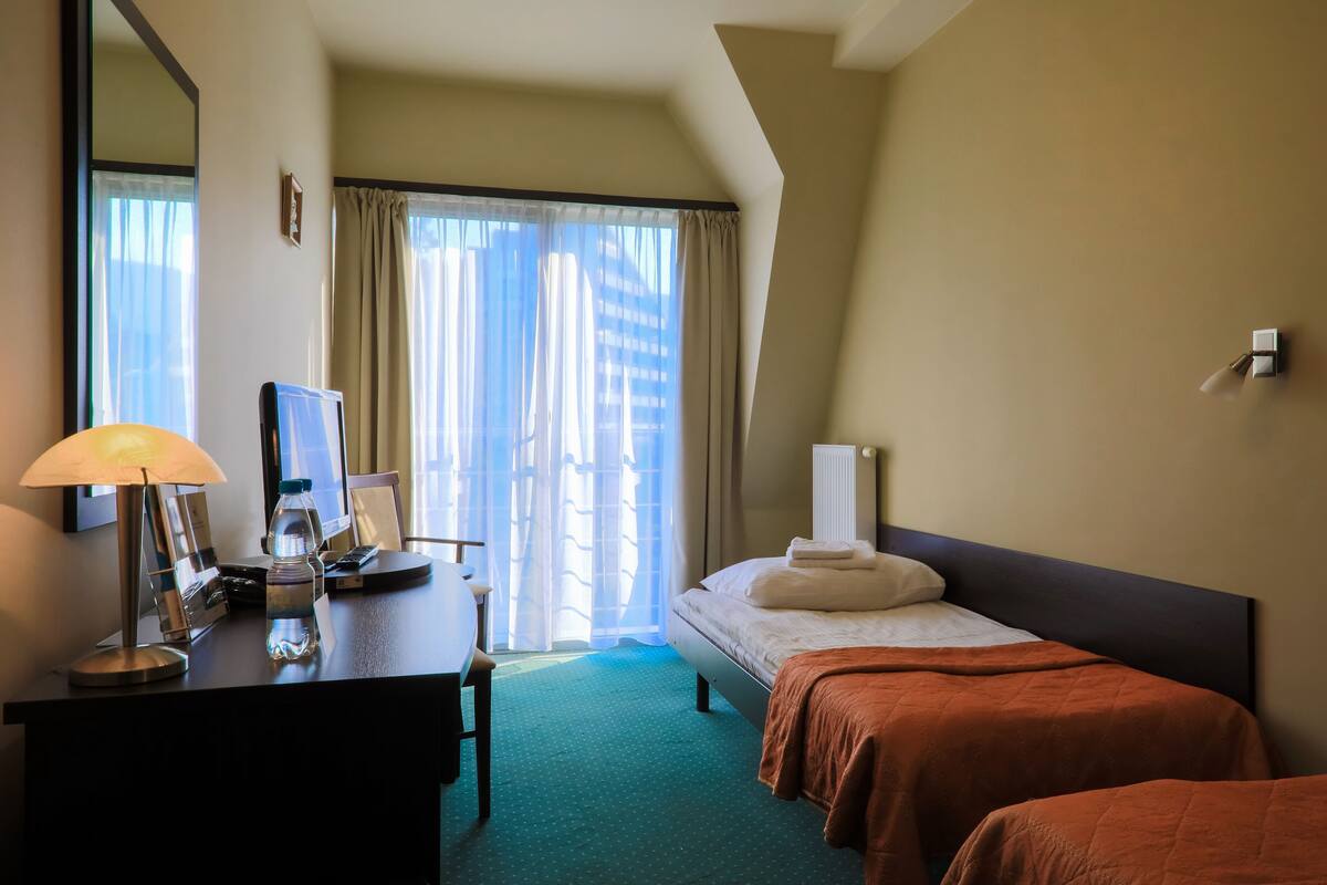 Habitación de hotel con escritorio, lámpara, botella de agua, cama y vistas de edificios a través del ventanal.