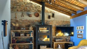 A cozy living room with a stone wall, a fireplace, a television, and a yellow sofa. - Madre del Agjua Rural House. Finca El Robledillo. (MONESTERIO)