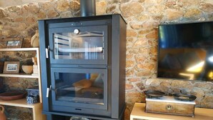 A wood stove with two glass doors, a woven basket, and a stone wall in the background. - Madre del Agjua Rural House. Finca El Robledillo. (MONESTERIO)
