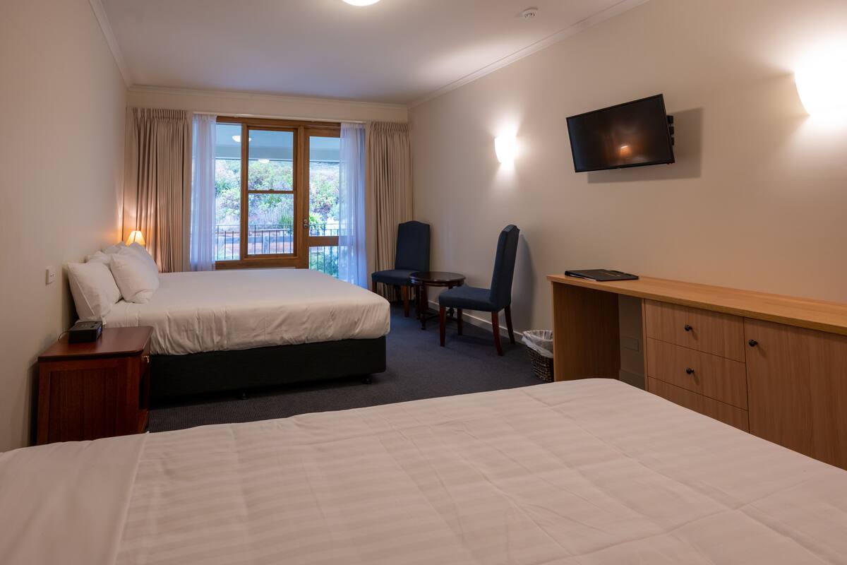 Ein Hotelzimmer mit zwei Betten, einem Schreibtisch, einem Stuhl, einem Fernseher und einem Fenster mit Vorhängen.